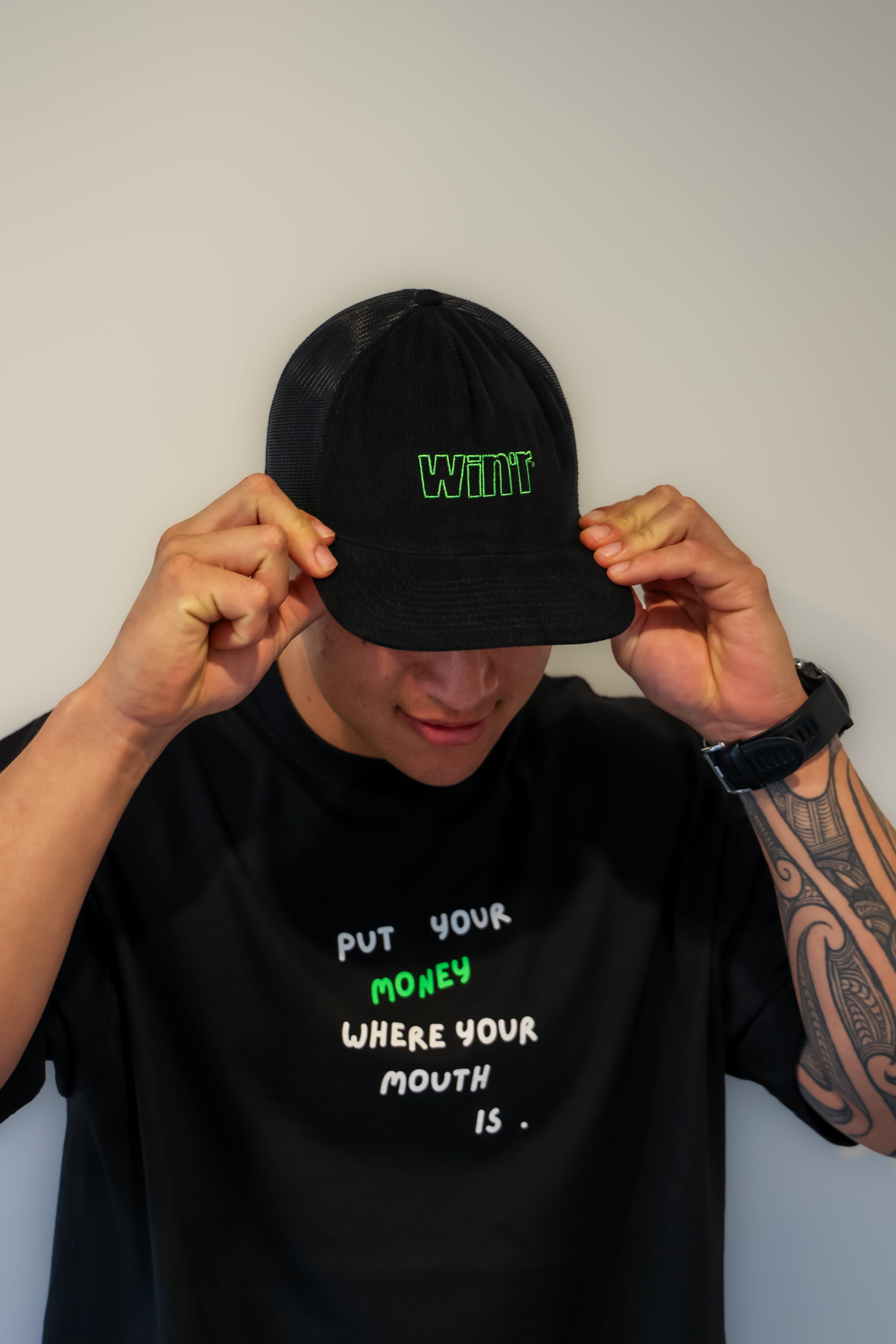 Person wearing a black Winr corduroy snapback cap with green embroidered text, paired with a black t-shirt with green and white graphic, against a plain background.