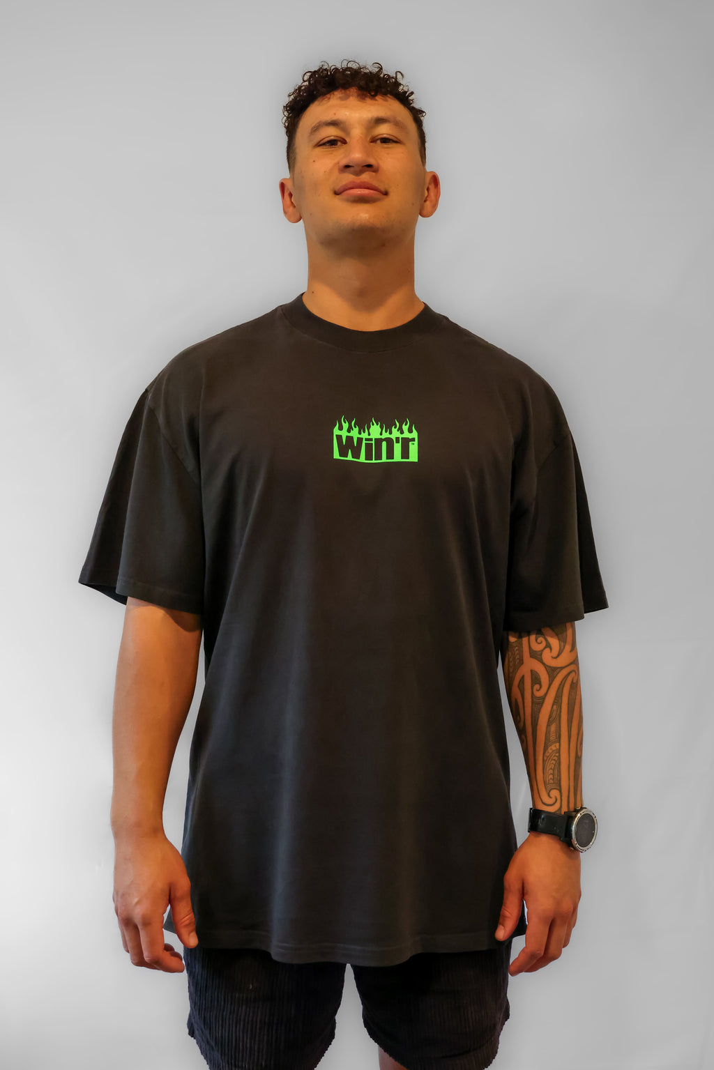 Person wearing a faded black Winr t-shirt with a green flame logo on the front, heavyweight cotton streetwear tee against a grey background.