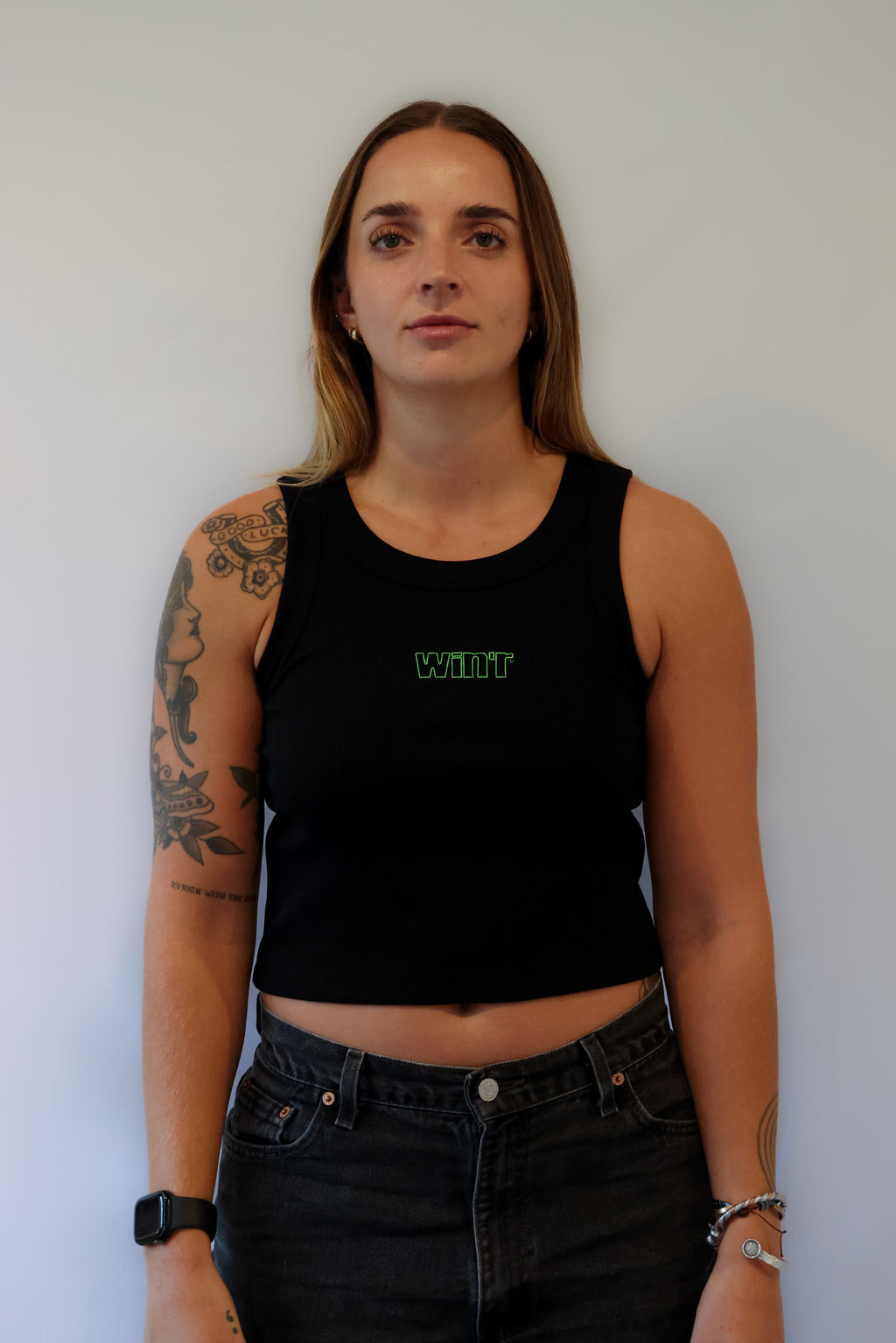 Woman wearing a black cotton Winr crop top with green embroidered text, fitted streetwear tank against a grey background.